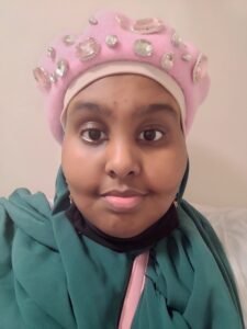 A Black woman smiling while wearing a green hijab, pink beret with gems and a white shirt with a pink strap from her bag being seen in the portrait.